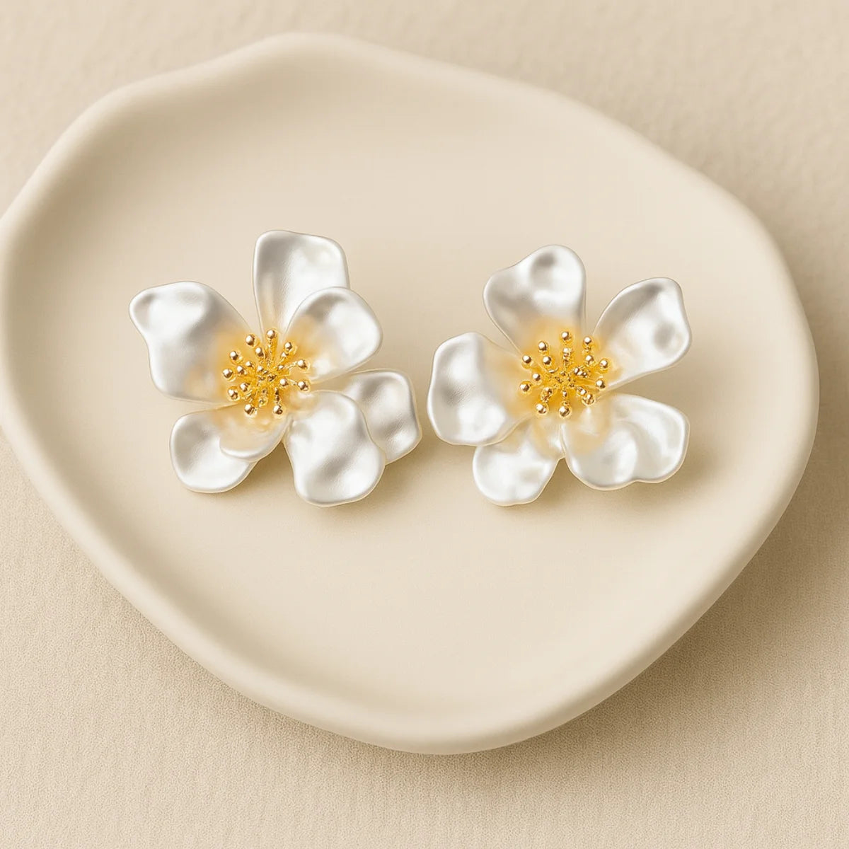 reate an ultra-realistic 1200×1200 px jewelry product image featuring the exact same white flower stud earrings from the reference image. The design must remain 100% unchanged: five soft-curved satin-finish petals per flower smooth pearlescent white texture gold stamen cluster in the center with rounded bead tips natural organic petal shape and proportions Do not change the shape, petal count, texture, size, or color. Earring Orientation: Both earrings must face straight toward the camera, upright and cent