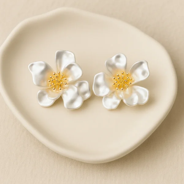 reate an ultra-realistic 1200×1200 px jewelry product image featuring the exact same white flower stud earrings from the reference image. The design must remain 100% unchanged: five soft-curved satin-finish petals per flower smooth pearlescent white texture gold stamen cluster in the center with rounded bead tips natural organic petal shape and proportions Do not change the shape, petal count, texture, size, or color. Earring Orientation: Both earrings must face straight toward the camera, upright and centered. No tilt, angle shift, rotation, or perspective distortion. Symmetrical spacing between both earrings. Background & Surface: Pure white background (#FFFFFF) Clean studio environment — no dish, no props, no beige tones, no textures. Completely plain and catalog-style. Lighting & Material Rendering: Soft diffused studio lighting. Natural pearlescent highlights on each petal to show curvature. Gentle warm reflections on the gold stamen beads. High clarity, no blown-out highlights. Shadow: Very soft, subtle, diffused shadow directly beneath each earring. Shadow must be light and realistic, like premium jewelry photography. Rendering Style: Hyper-realistic, luxury brand quality. Crisp edges, balanced contrast. Zero artificial or plastic look. Negative Prompt: no design changes, no extra petals, no dish, no beige background, no props, no blur, no noise, no reflections of objects, no warped shapes, no tilt, no 3/4 angle, no overly matte finish, no overly glossy finish, no AI artifacts.